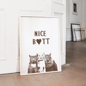 May include: A white framed poster with the text "NICE B❤TT" in brown lettering. Three cats are pictured below the text, with the cat in the middle looking directly at the viewer.