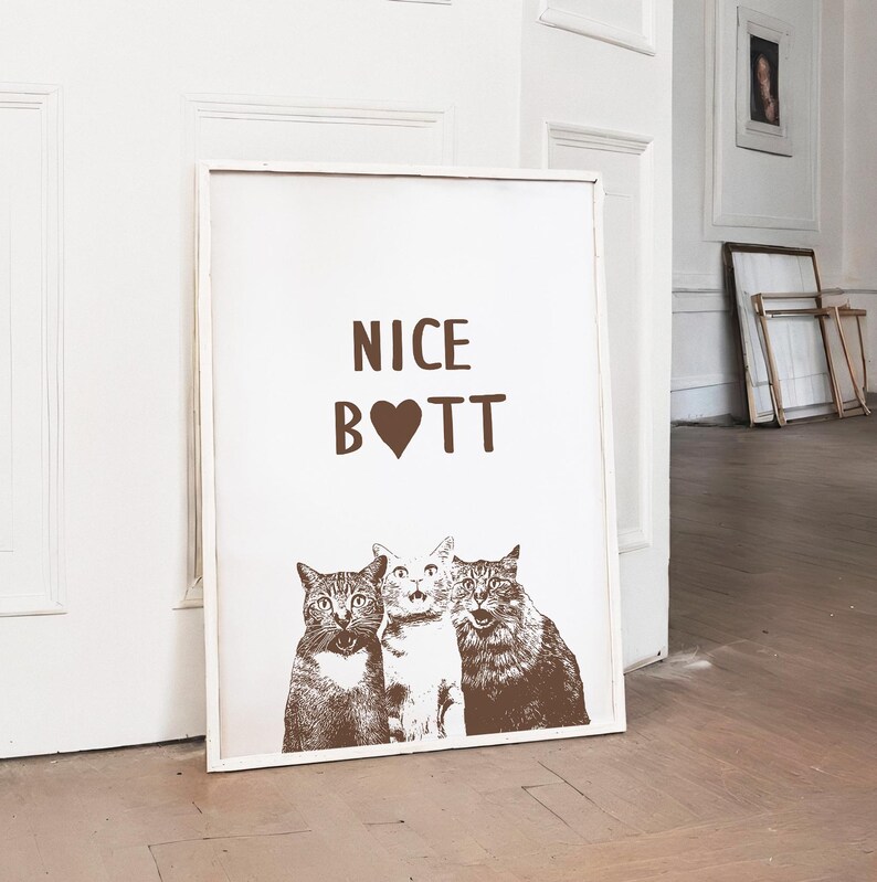 May include: A white framed poster with the text "NICE B❤TT" in brown lettering. Three cats are pictured below the text, with the cat in the middle looking directly at the viewer.