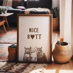 May include: A framed print with the text "NICE B♡TT" and a picture of three cats. The frame is brown wood with a decorative border.
