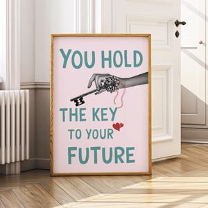 Op de afbeelding: Een ingelijste print met de tekst "YOU HOLD THE KEY TO YOUR FUTURE" in teal op een roze achtergrond. De afbeelding toont een hand die een sleutel vasthoudt met een rood hart en een sleutelontwerp.