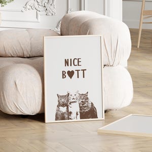 May include: A white framed print with the text "NICE BOTT" in brown lettering with a heart in place of the "O". Three cats are pictured below the text, two with their heads turned to the left and one with its head turned to the right.