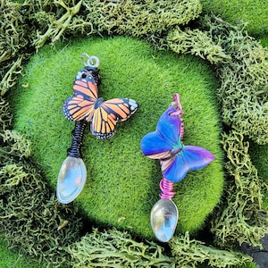 May include: Two spoons with butterfly designs. One spoon has a black wire wrap and a monarch butterfly design. The other spoon has a pink wire wrap and a blue butterfly design. Both spoons are on a bed of green moss.