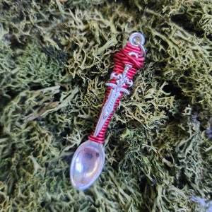 May include: A silver spoon with a red wire-wrapped handle shaped like a sword. The spoon is lying on a bed of green moss.