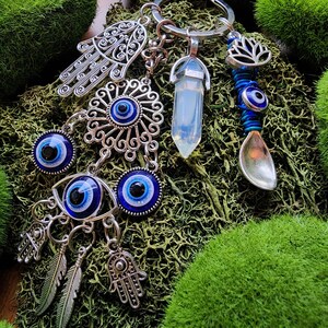 May include: A silver keychain with various charms, including an eye-shaped pendant, a crystal, a lotus flower, and a spoon-shaped charm. The charms are arranged on a bed of green moss.