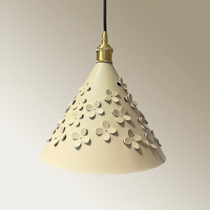 Puede incluir: Lámpara colgante de color crema con una pantalla cónica adornada con numerosos recortes de flores tridimensionales. La lámpara tiene un casquillo de color latón y un cable negro. Diseño elegante y decorativo para iluminación interior.