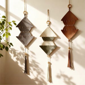 May include: Three leather wall hangings in shades of gray, cream, and brown, each with a geometric design. The pieces are suspended by twine and feature wooden beads and tassels. The wall decor is displayed against a white wall.