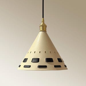 May include: Cream-colored pendant light with a conical shade. The shade features black rectangular accents and small rectangular cutouts near the top. The light fixture has a brass-colored top and a black cord.