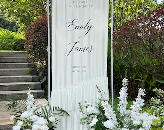 May include: A white fabric wedding welcome sign on a metal frame featuring the text Welcome to our wedding, Emily + James, Sydney, SYD, and 09.30.2026. The sign has a ruffled bottom section and is displayed outdoors with white floral arrangements.