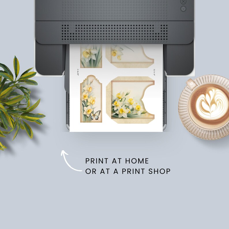 May include: A printer printing a sheet with floral designs, including daffodils and a butterfly. The sheet has cut-out shapes for tags and other crafts. Text on the image reads "PRINT AT HOME OR AT A PRINT SHOP." A cup of coffee sits nearby.