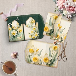 May include: A collection of stationery items with daffodil illustrations. Includes a dark green card with pockets, several cards, a spool of ribbon, scissors, and a cup of tea. The daffodils have yellow centres and white petals.