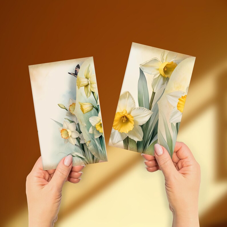 May include: Two rectangular cards with watercolour illustrations of daffodils. The cards show white petals with yellow centres and green leaves. One card has a butterfly. The background is a warm, neutral tone.