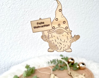 May include: Wooden Christmas gnome ornament with a dotted hat and a sign that reads "Frohe Weihnachten". The gnome is hanging from a string. Other wooden ornaments are on a wooden tray with greenery and gold ornaments.