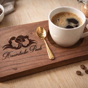 Könnte beinhalten: Eine weiße Kaffeetasse mit Kaffee steht auf einem Holztablett mit einem goldenen Löffel. Das Tablett hat ein geschnitztes Design und den Text "Haarstudio Pauli". Kaffeebohnen befinden sich in einer weißen Schüssel.