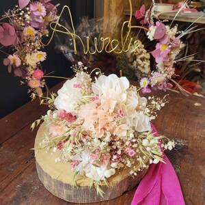 May include: A dried flower arrangement with a gold wire hoop with the name "Aurelia" on it. The flowers are in shades of pink, white, and green. The arrangement is on a wooden slice and a pink fabric is on the table.