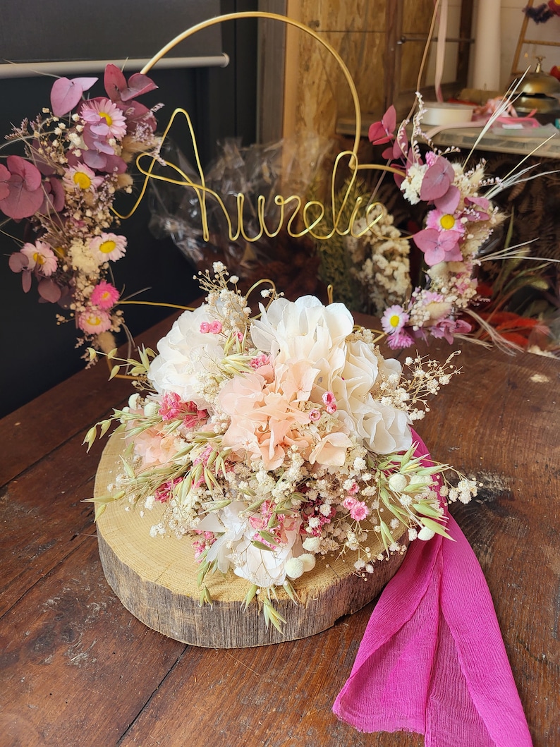 May include: A dried flower arrangement with a gold wire hoop with the name "Aurelia" on it. The flowers are in shades of pink, white, and green. The arrangement is on a wooden slice and a pink fabric is on the table.
