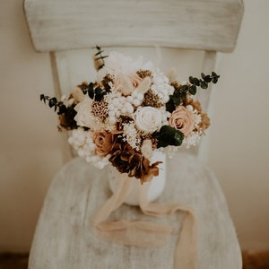 May include: A bouquet of dried flowers in shades of white, brown, and pink, arranged in a white vase with a beige ribbon. The bouquet is sitting on a white wooden chair.