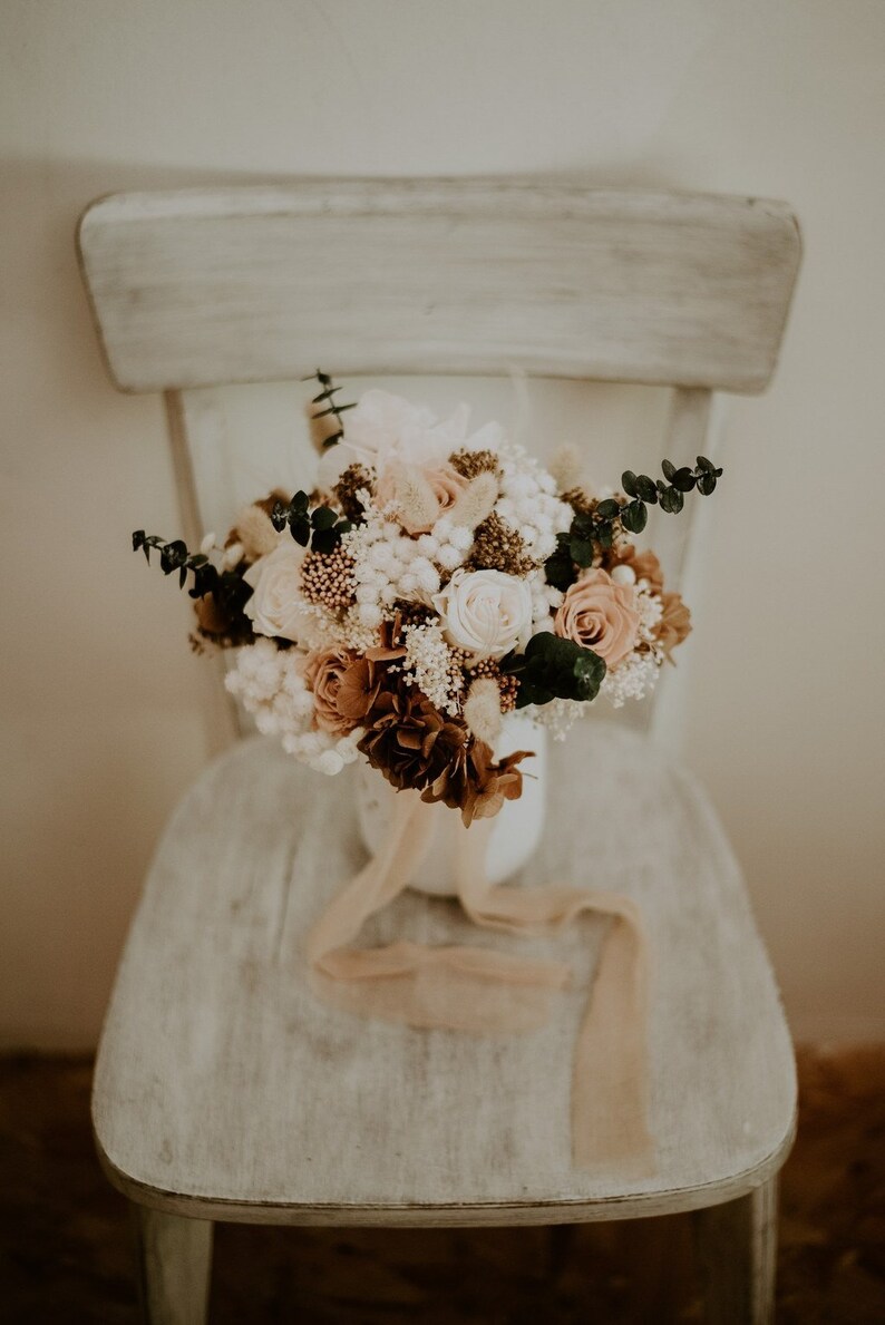 May include: A bouquet of dried flowers in shades of white, brown, and pink, arranged in a white vase with a beige ribbon. The bouquet is sitting on a white wooden chair.