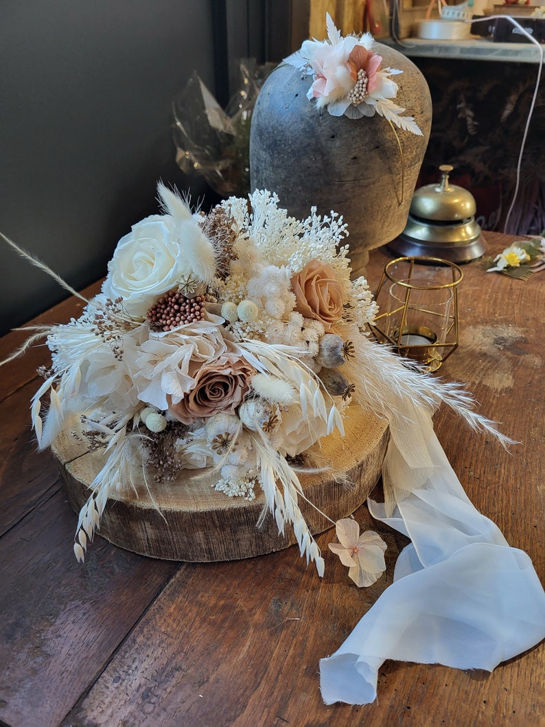 May include: A dried flower bouquet with white and beige flowers, brown accents, and long white grasses. The bouquet is arranged on a wooden slice and a white sheer ribbon is draped over the arrangement.