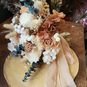 May include: A dried flower bouquet with white, beige, and brown flowers, eucalyptus, and dried grasses. The bouquet is tied with a beige ribbon and sits on a wooden slice.