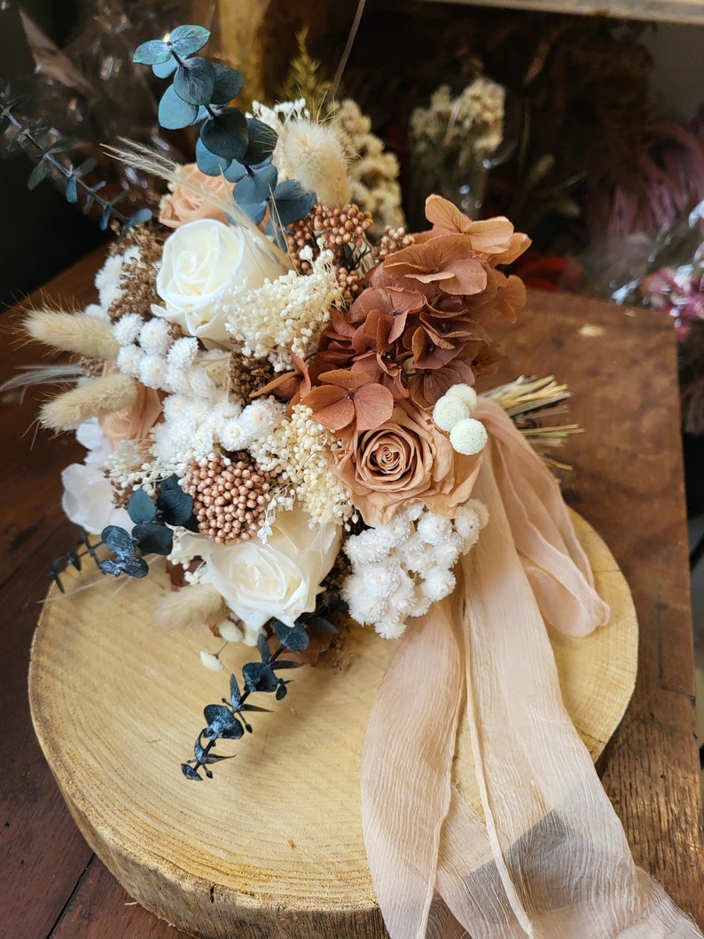 May include: A dried flower bouquet with white, beige, and brown flowers, eucalyptus, and dried grasses. The bouquet is tied with a beige ribbon and sits on a wooden slice.