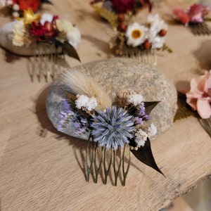 May include: A gold hair comb decorated with dried flowers, including a blue thistle, white flowers, and a brown dried leaf. The comb is resting on a grey rock.