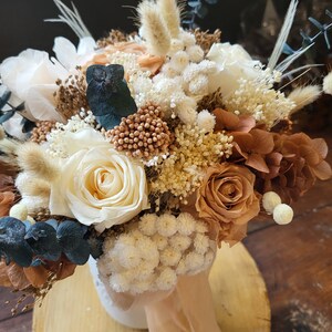 May include: A dried flower arrangement in a white vase with a beige ribbon. The bouquet features white, beige, and brown roses, white baby's breath, and dried grasses.