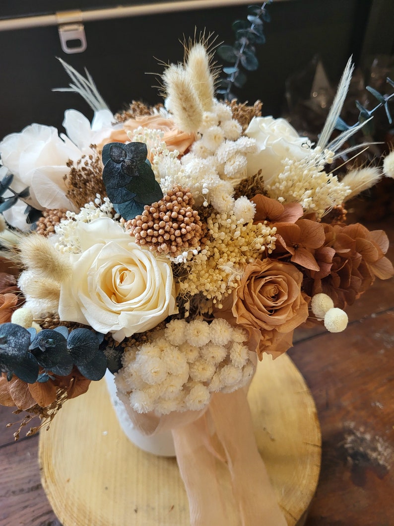 May include: A dried flower arrangement in a white vase with a beige ribbon. The bouquet features white, beige, and brown roses, white baby's breath, and dried grasses.