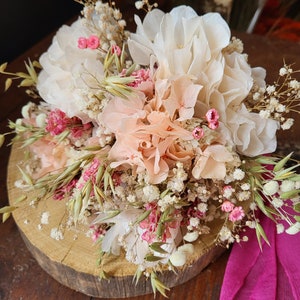 May include: A dried flower bouquet with white, peach, and pink flowers, and green and brown grasses. The bouquet is arranged on a wooden slice and a pink fabric is in the foreground.