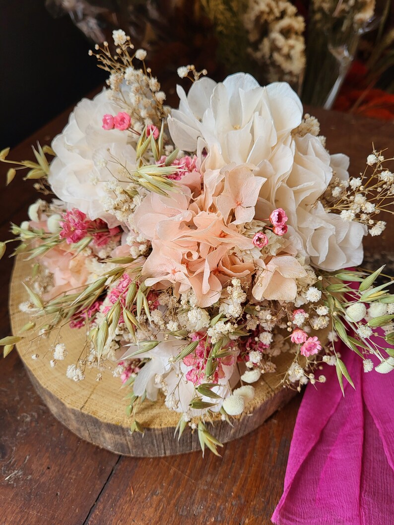 May include: A dried flower bouquet with white, peach, and pink flowers, and green and brown grasses. The bouquet is arranged on a wooden slice and a pink fabric is in the foreground.