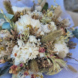 May include: A dried flower bouquet with white, cream, and brown flowers, dried grasses, and seed pods. The bouquet is arranged in a white paper wrapping.