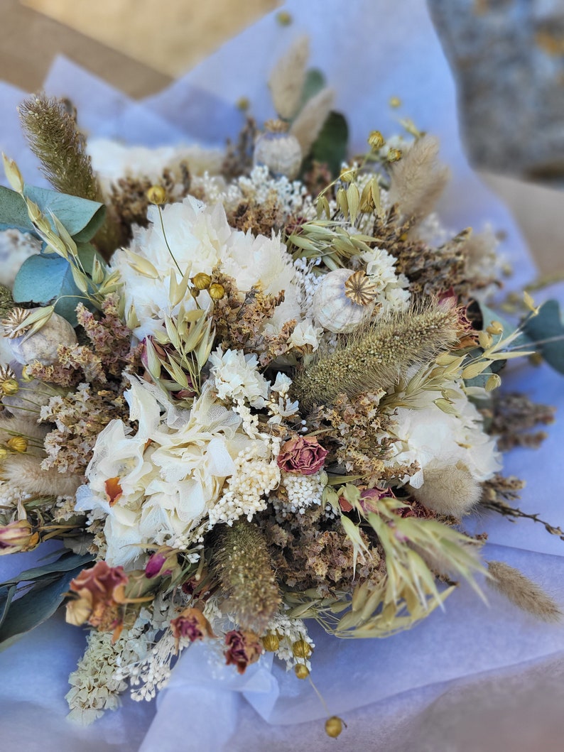 May include: A dried flower bouquet with white, cream, and brown flowers, dried grasses, and seed pods. The bouquet is arranged in a white paper wrapping.