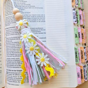 May include: A handmade bookmark tassel with daisy accents. The tassel features white, pink, yellow, and green ribbons and threads, with wooden beads at the top. The bookmark is placed on an open book with labeled tabs.
