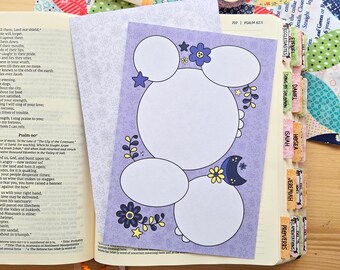 May include: A light purple card with four blank white circular spaces for writing, decorated with yellow and blue flowers, stars, and a crescent moon. The card rests on an open Bible with visible text and colorful book tabs on the side.