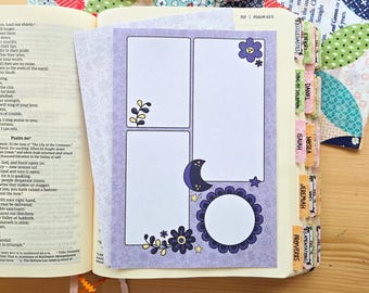 May include: A purple paper insert with three white rectangular and circular frames, decorated with dark purple flowers, a crescent moon, and small yellow stars, placed inside an open Bible with labeled book tabs.