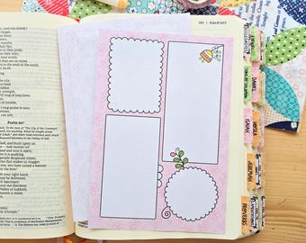 May include: A pink paper insert with three blank white frames featuring scalloped and smooth edges, decorated with small floral line art, placed inside an open Bible with colorful book tabs labeled with various book names.