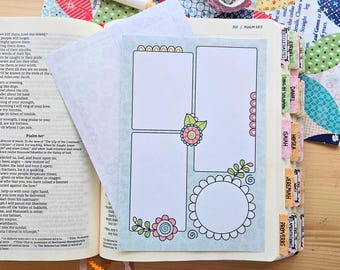 May include: A light blue paper insert with three blank rectangular frames and one circular frame, decorated with pink and yellow flower illustrations, placed inside an open book with printed text and colorful index tabs on the side.