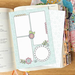 May include: A light blue card with a floral pattern featuring two rectangular frames, one circular frame, and decorative pink and green flower illustrations. The card is placed on an open book with visible text and colorful index tabs on the side.