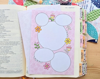 May include: A light pink paper insert with five blank white circular and oval spaces for writing, decorated with colorful flower illustrations, placed inside an open Bible with indexed tabs and visible text from Psalm 60 and 62.