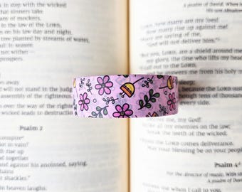 May include: A roll of light purple washi tape featuring a repeating pattern of small pink flowers with yellow centers, green leaves, and thin black stems. The tape is displayed horizontally against a background of printed book pages.