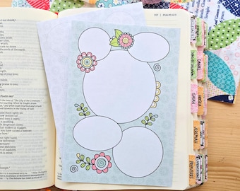 May include: A light blue paper insert with five blank circular spaces for notes, decorated with pink and yellow flower illustrations and green leaves, placed inside an open Bible with colorful book tabs and markers.