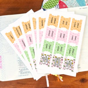 May include: A set of adhesive Bible index tabs printed on white paper with scalloped edges. The tabs feature pastel floral patterns in yellow, pink, and green, with black text identifying various books of the Bible such as Genesis, Ruth, and Numbers.
