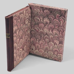 Może przedstawiać: Otwarta książka z bordowym grzbietem i okładką. Strony i okładka mają dekoracyjny wzór w odcieniach bordowego, kremowego i brązowego. Grzbiet ma złote litery. Książka jest na jasnoszarym tle.
