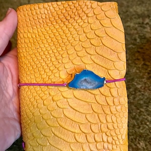 May include: A yellow journal with a textured, scale-like pattern. A blue and silver gemstone is attached with a pink elastic band. The journal is held in a hand.