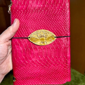 May include: A hot pink, textured leather journal with a gold-colored bee-shaped clasp. The journal is closed with a black elastic band. A small gold charm hangs from the top left corner.