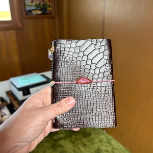 May include: A dark brown leather journal with a textured, crocodile-like pattern. The journal is secured with a pink elastic band and a red stone accent. A decorative charm hangs from the top. The journal is held in a person's hand.