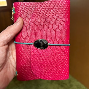 May include: A hot pink, textured leather journal with a black stone accent and teal elastic closure. The journal has a snake-skin pattern and gold-colored hardware. The journal is closed and held by a hand.