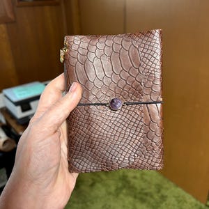 May include: Brown leather journal with a textured, reptile-like pattern. The journal is secured with a black elastic band and a purple gemstone button. A gold-colored charm hangs from the top.