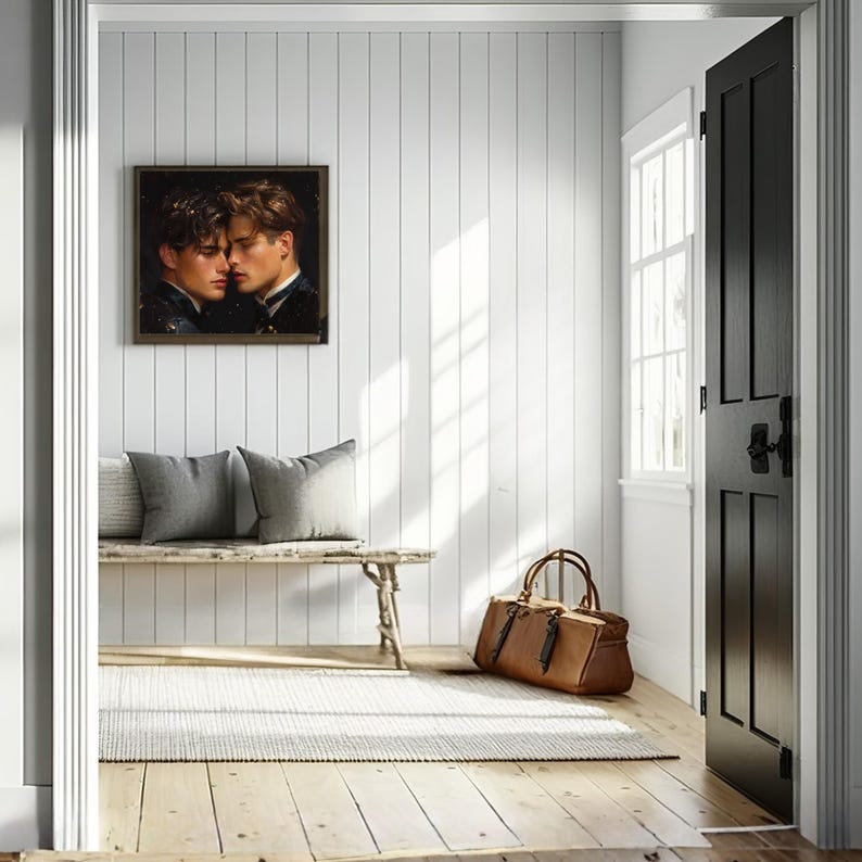 May include: A framed wall art piece depicting two individuals in close proximity, mounted on a white vertical panelled wall above a wooden bench with two grey pillows, a tan leather duffel bag, and a textured rug on a light wood floor.