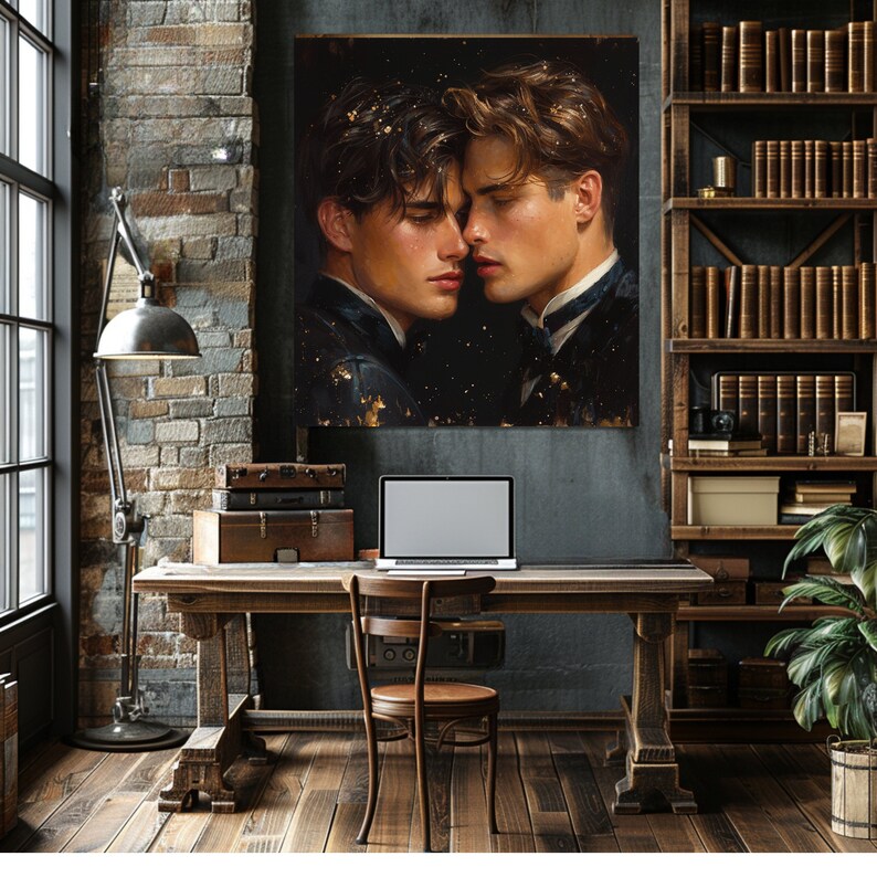 May include: A square wall art print featuring a portrait of two individuals with dark hair facing each other closely against a dark background with gold flecks. The artwork is displayed above a wooden desk with a laptop and stacked vintage suitcases.