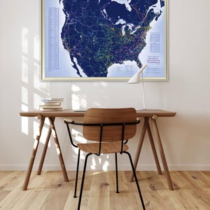 May include: A framed map of North America, showcasing electricity generation and transmission lines. The map is predominantly blue with colorful lines representing the electrical grid. The frame is silver, and the map hangs above a wooden desk with a chair.